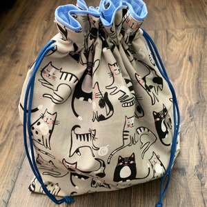 May include: A gray drawstring bag with a repeating pattern of cartoon cats in various poses. The cats are black, white, and gray with pink accents. The bag has a blue lining and a blue drawstring.