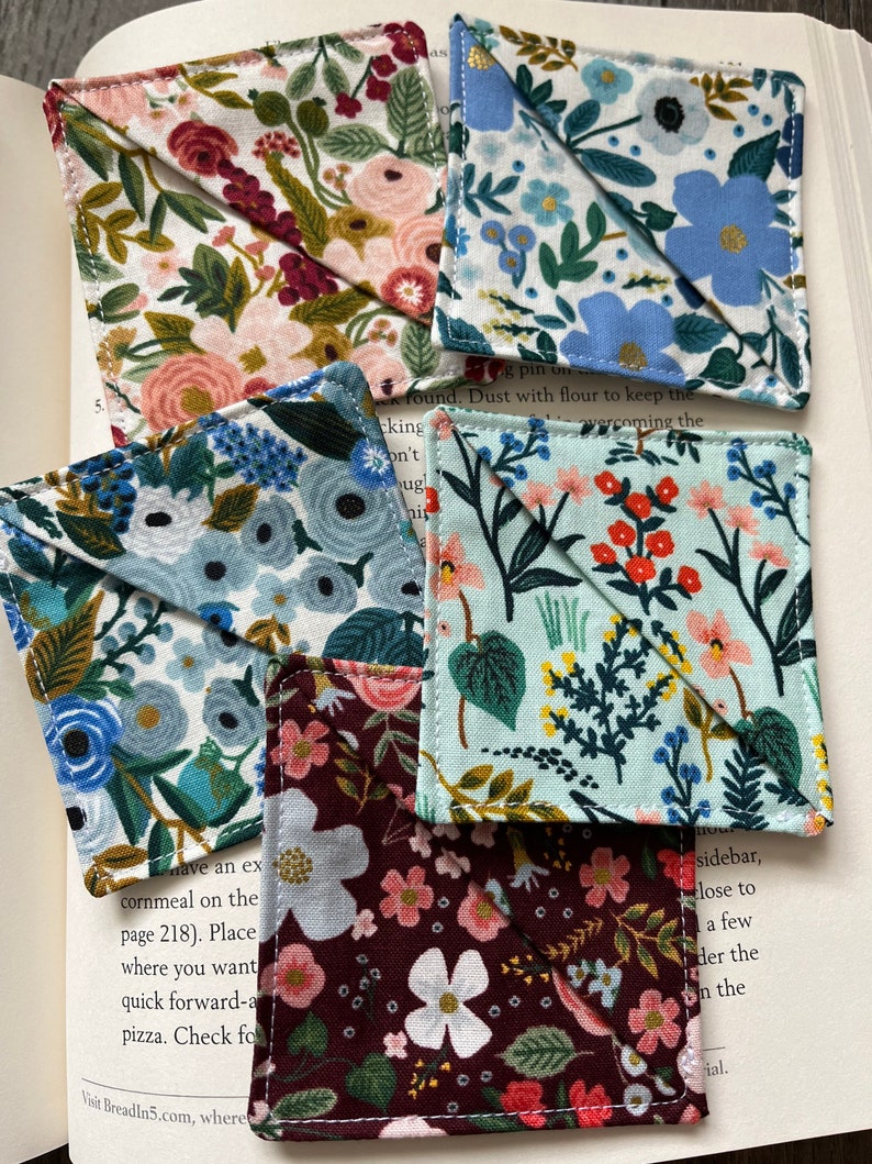 May include: Four fabric bookmarks with floral patterns. The bookmarks are folded into a triangle shape. The patterns include red, pink, blue, and green flowers on a white background.