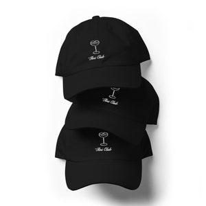 May include: Three black baseball caps stacked on top of each other. Each cap features a white embroidered design of a cocktail glass and the text "Tini Club".