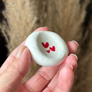 Può includere: Un piccolo oggetto rotondo in ceramica bianca con un centro concavo caratterizzato da tre cuori rossi dipinti a mano. La superficie ha una finitura opaca e leggermente ruvida, e l'oggetto è tenuto in mano per mostrare il disegno.