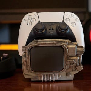 May include: A white and black PlayStation 5 controller rests on a detailed, weathered-looking stand. The stand has a rectangular screen and a textured, metallic appearance. The controller's buttons and joysticks are visible.