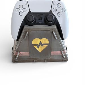 May include: A white and black PlayStation 5 controller rests on a custom-designed charging stand. The stand is gray with a gold heart and heartbeat design. The stand has a metallic finish and a pink accent.