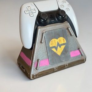 May include: A white PlayStation 5 controller rests on a custom-designed stand. The stand is gray with pink stripes and a yellow broken heart graphic. The controller is white with black accents.