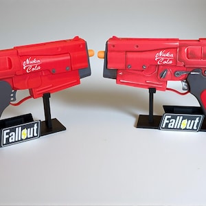 May include: Two red toy guns with the words "Nuka Cola" printed on the side. The guns have gray grips and are displayed on black stands with "Fallout" printed on them. The background is white.