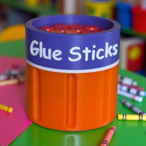 May include: A cylindrical container for glue sticks, featuring a vibrant design. The top section is a deep purple, with the words "Glue Sticks" in white. The lower portion is bright orange. The container is filled with glue sticks.
