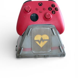 May include: A red Xbox controller resting on a gray and red base. The base features a gold heart and heartbeat design. The controller has black buttons and joysticks.