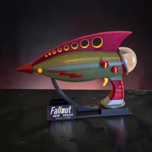 May include: A replica of the Euclid C-Finder weapon from Fallout New Vegas, featuring an olive green body, maroon accents, a yellow trigger, and a clear dome. It rests on a black stand with a plaque reading Fallout New Vegas Euclid C-Finder.