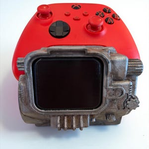 May include: A red Xbox controller with black buttons and joysticks is paired with a metallic, industrial-style device. The device has a rectangular black screen and a weathered, textured appearance, suggesting a vintage or post-apocalyptic aesthetic.
