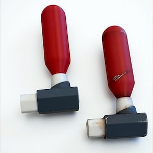 May include: Two red and gray bottle stoppers with a rectangular base and a cylindrical top. The stoppers have a white section between the red top and the gray base. One stopper has a scratch.