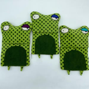 May include: Three green frog-shaped plush toys with polka dots. Each frog has a different coloured eye. The frogs are made of soft, fuzzy fabric.