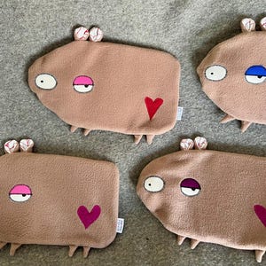 May include: Four light brown plush creatures with quirky features, including different coloured eyes and heart shapes. Each has small feet and patterned ears. They are arranged on a grey surface. The creatures are approximately 20 cm long.