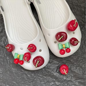 May include: A pair of off-white slip-on clogs featuring multiple red cherry charms and light green bows attached to the top surface. The shoes have a smooth, molded construction with ventilation holes and a single loose cherry charm nearby.