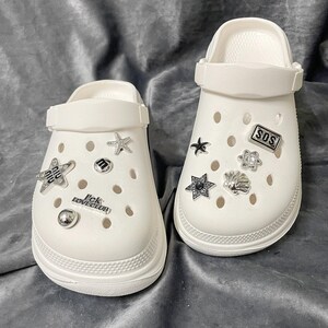May include: A pair of white clogs with decorative charms. The charms include stars, a seashell, and the letters "M" and "SOS". The text "I'ck LOVECLUB" is also visible. The clogs have a thick sole and adjustable strap.