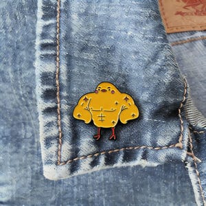 May include: A yellow enamel pin depicting a strong cartoon chick with red feet. The pin is set against a blue denim jacket, highlighting its bright colours and playful design. This accessory adds a touch of humour and personality.