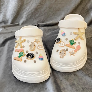 Könnte beinhalten: Weiße Clogs, verziert mit verschiedenen Charms, darunter Seesterne, Muscheln und Eiswaffeln. Die Charms sind in Gold, Schwarz, Rosa und Grün gehalten. Die Clogs haben eine dicke Sohle und einen Riemen.