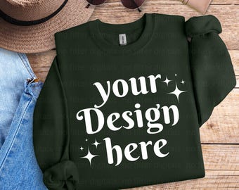 May include: A dark green sweatshirt with the text "your Design here" in white. The sweatshirt is laid flat on a wooden surface with a pair of sunglasses, a hat, and a purse.