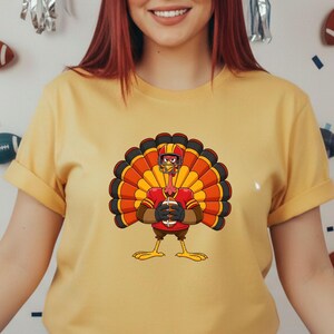 Op de afbeelding: Een geel T-shirt met een cartoon kalkoen in een voetbaluniform. De kalkoen houdt een voetbal vast. De achtergrond bevat voetbaldecoraties en posters voor Super Bowl LXIX.