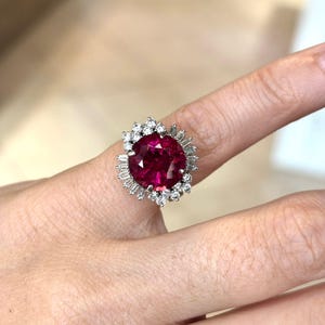 May include: A large, oval-cut ruby ring with a halo of small, round diamonds and baguette-cut diamonds. The ruby is a deep, vibrant red color, and the ring is set in a silver or white gold band. The ring is on a finger.
