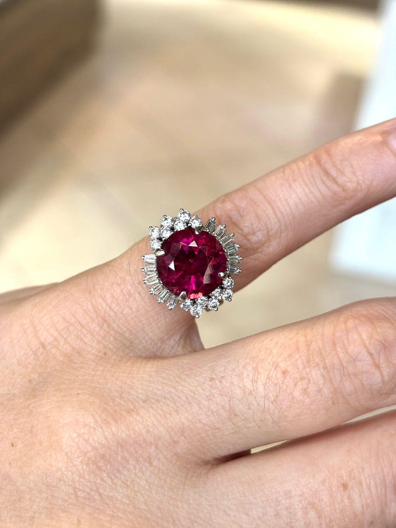 May include: A large, oval-cut ruby ring with a halo of small, round diamonds and baguette-cut diamonds. The ruby is a deep, vibrant red color, and the ring is set in a silver or white gold band. The ring is on a finger.