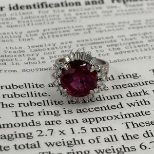 May include: A silver ring featuring a large, oval-cut, deep red rubellite gemstone, accented with smaller diamonds. The ring's design includes a halo of diamonds around the central stone. The ring weighs 6.7 grams.