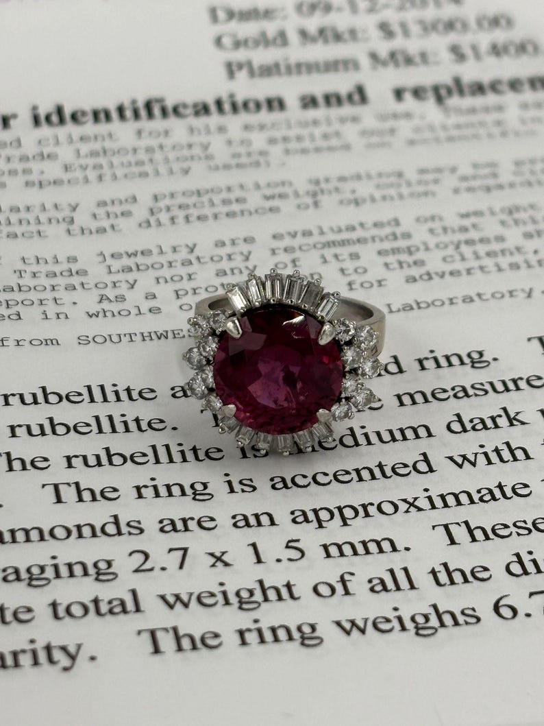 May include: A silver ring featuring a large, oval-cut, deep red rubellite gemstone, accented with smaller diamonds. The ring's design includes a halo of diamonds around the central stone. The ring weighs 6.7 grams.