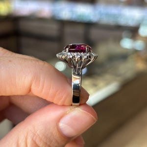 May include: A silver ring featuring a large, oval-cut, deep red gemstone surrounded by a halo of smaller, clear gemstones. The ring's band is a polished silver. The setting is intricate, with the gemstones held in place by delicate prongs.