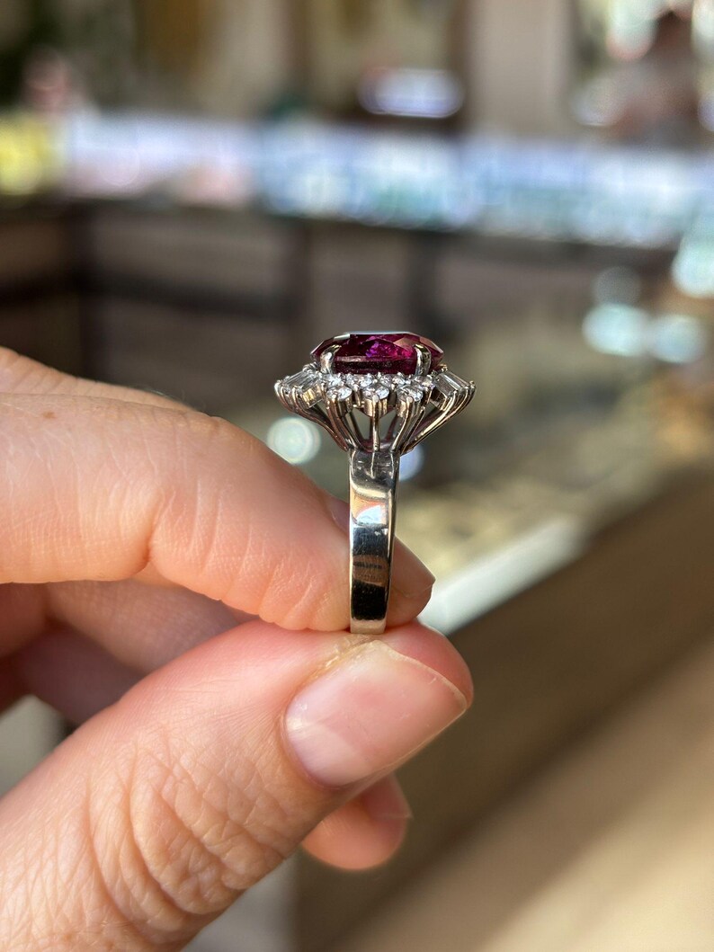 May include: A silver ring featuring a large, oval-cut, deep red gemstone surrounded by a halo of smaller, clear gemstones. The ring's band is a polished silver. The setting is intricate, with the gemstones held in place by delicate prongs.