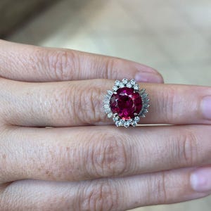 May include: A large, round, ruby-colored gemstone ring with a halo of smaller, clear gemstones. The ring's setting is silver-colored, and the gemstones are arranged in a cluster around the central ruby. The ring is on a person's finger.