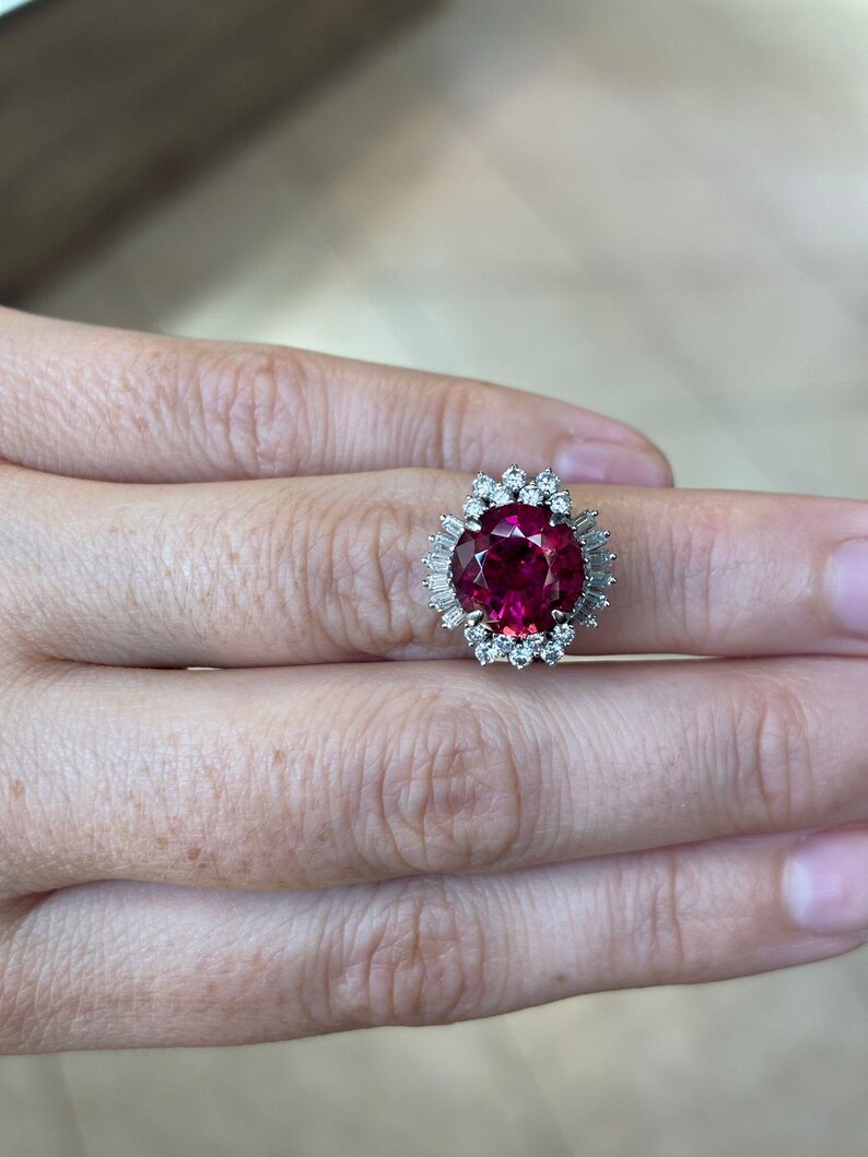 May include: A large, round, ruby-colored gemstone ring with a halo of smaller, clear gemstones. The ring's setting is silver-colored, and the gemstones are arranged in a cluster around the central ruby. The ring is on a person's finger.