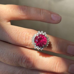 May include: A silver ring featuring a large, oval-cut, deep red gemstone surrounded by smaller, clear gemstones. The ring's design includes a halo of baguette-cut stones around the central stone, creating a sparkling effect. The ring is displayed on a finger.