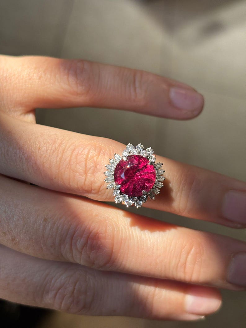May include: A silver ring featuring a large, oval-cut, deep red gemstone surrounded by smaller, clear gemstones. The ring's design includes a halo of baguette-cut stones around the central stone, creating a sparkling effect. The ring is displayed on a finger.
