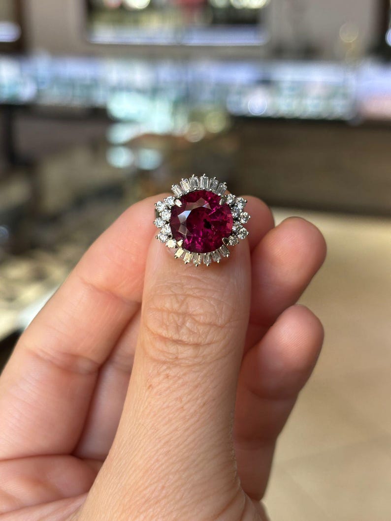 May include: A close-up of a ring featuring a large, round, deep red gemstone. The gemstone is surrounded by smaller, clear, baguette-cut and round-cut stones set in a silver or white gold band. The ring is held up by a person's fingers.