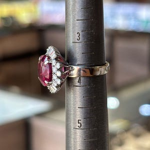 May include: A vintage ring with a large, oval-cut, deep pink gemstone as the centerpiece. The ring is set with smaller, clear gemstones and is on a ring sizer. The ring's band is a light gold color.