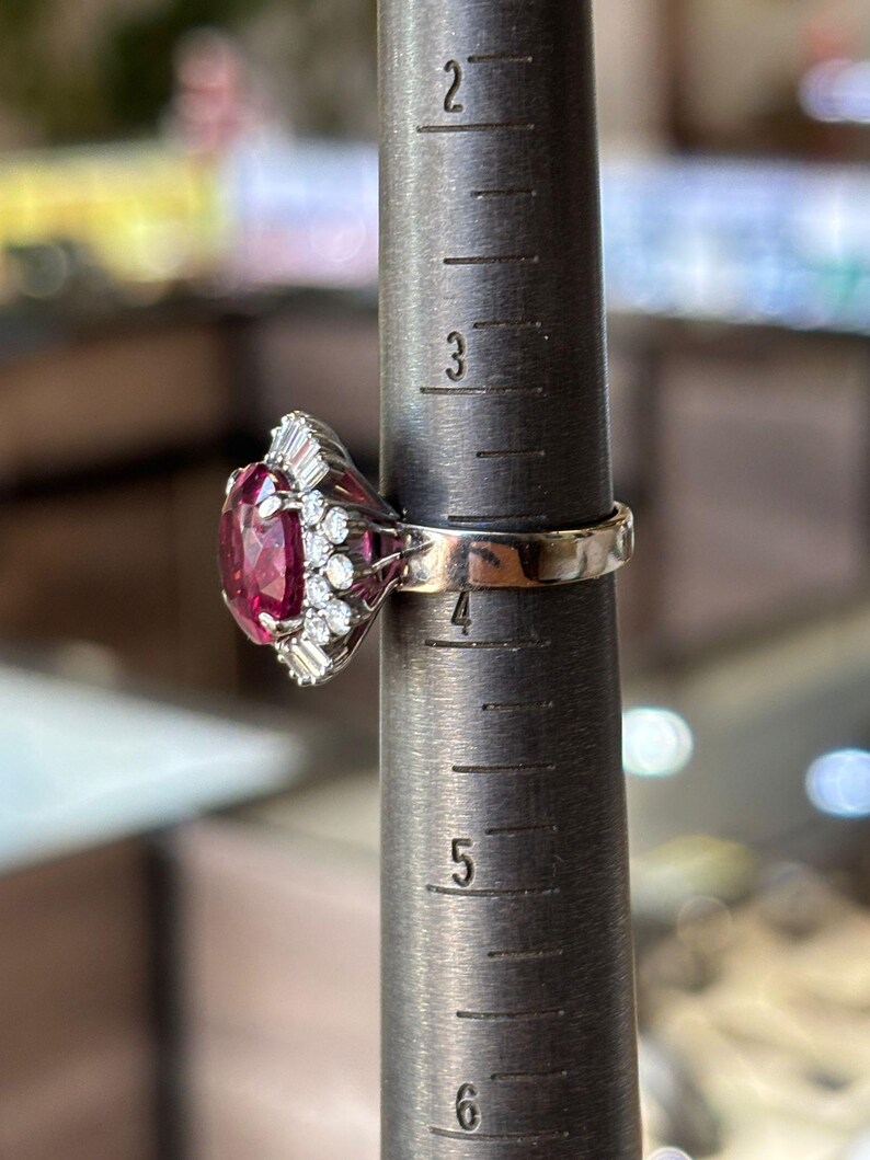 May include: A vintage ring with a large, oval-cut, deep pink gemstone as the centerpiece. The ring is set with smaller, clear gemstones and is on a ring sizer. The ring's band is a light gold color.