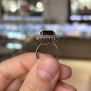 May include: A silver ring featuring a large, oval-cut, deep red gemstone surrounded by a halo of smaller, clear stones. The ring's band is thin and polished, and the setting has a detailed, vintage-inspired design. The ring is held up against a blurred background.