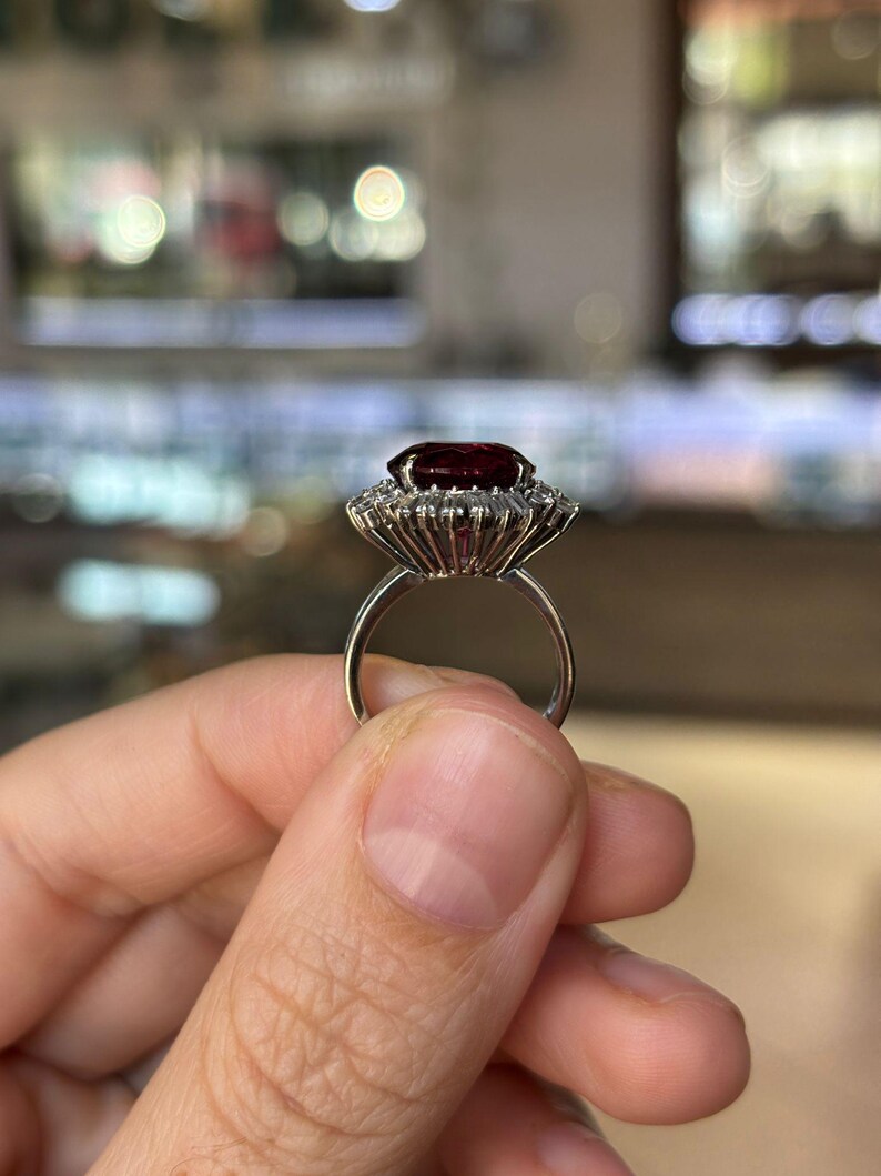 May include: A silver ring featuring a large, oval-cut, deep red gemstone surrounded by a halo of smaller, clear stones. The ring's band is thin and polished, and the setting has a detailed, vintage-inspired design. The ring is held up against a blurred background.