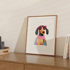K&ouml;nnte beinhalten: Eine gerahmte Illustration eines schwarz-wei&szlig;en Hundes, der eine rosa Sonnenbrille und einen bunten Pullover tr&auml;gt. Der Hund schaut direkt auf den Betrachter.