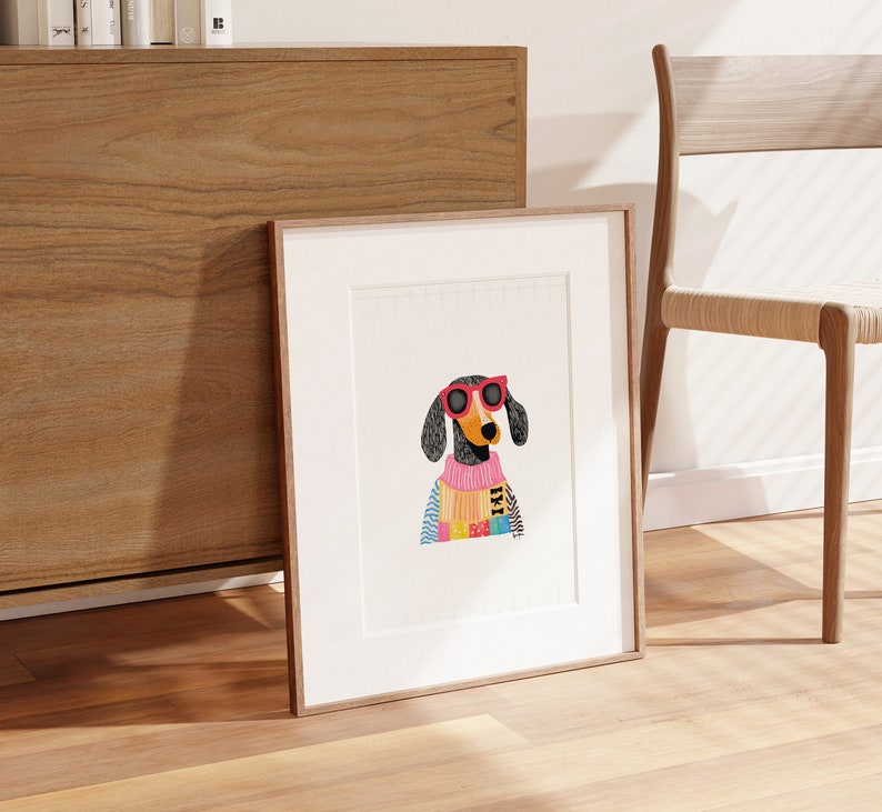K&ouml;nnte beinhalten: Eine gerahmte Illustration eines Dackels mit rosa Sonnenbrille und einem bunten Pullover. Der Hund schaut direkt auf den Betrachter.