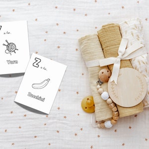 Op de afbeelding: Twee witte kaarten met zwarte letters en illustraties. De eerste kaart zegt "Y is voor... Garen" en heeft een afbeelding van een bol garen met breinaalden. De tweede kaart zegt "Z is voor... Courgette" en heeft een afbeelding van een courgette. De kaarten liggen op een witte stof met een bruin sterrenpatroon.