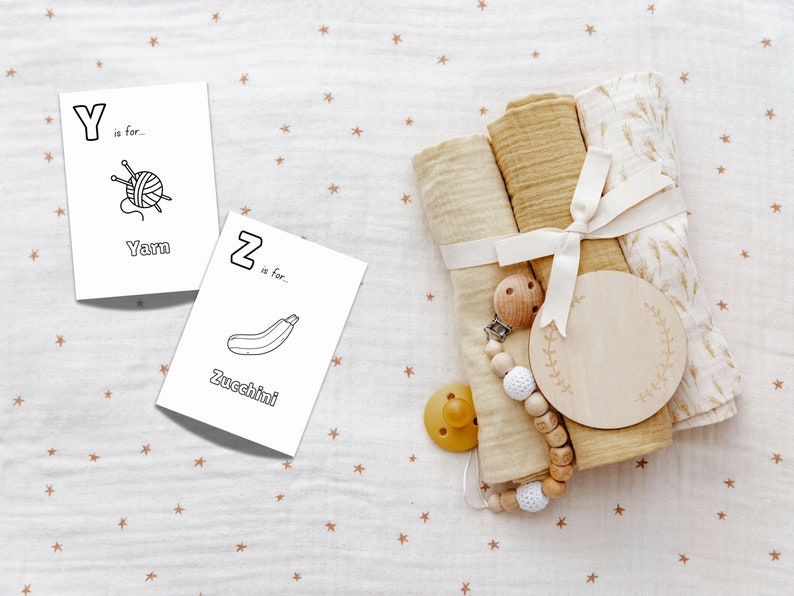 Op de afbeelding: Twee witte kaarten met zwarte letters en illustraties. De eerste kaart zegt "Y is voor... Garen" en heeft een afbeelding van een bol garen met breinaalden. De tweede kaart zegt "Z is voor... Courgette" en heeft een afbeelding van een courgette. De kaarten liggen op een witte stof met een bruin sterrenpatroon.