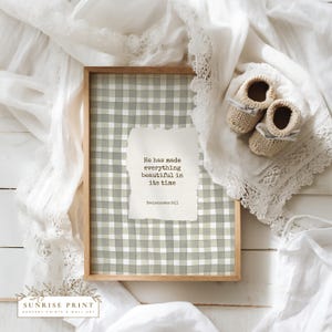 May include: A framed print with a green and white gingham pattern and the quote, "He has made everything beautiful in its time." The frame is light brown. Two small, beige knitted baby booties with gray bows rest on a white lace-trimmed cloth.