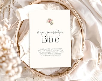 Op de afbeelding: Een crèmekleurig bord met de tekst "please sign our baby's Bible" in elegant schrift. Een bloemenkruisontwerp staat bovenaan. Het bord staat in een gevlochten mand met witte stof.