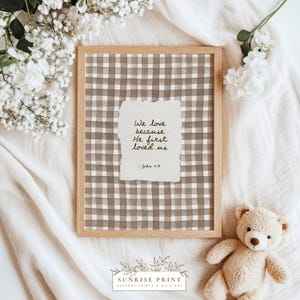 Könnte beinhalten: Ein gerahmter Druck mit hellbraunem Vichy-Muster und einem zerrissenen Papiereinsatz mit dem Text "We love because He first loved us." Der Rahmen ist aus hellem Holz. Ein Teddybär und weiße Blumen sind im Bild.
