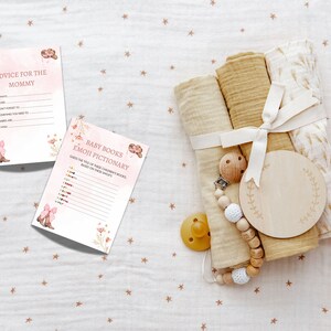 Peut inclure: Deux cartes de jeu de baby shower roses et blanches avec des motifs floraux. La premi&egrave;re carte dit "Conseils pour la maman" et contient une liste d'invites. La deuxi&egrave;me carte dit "Pictionary des livres pour b&eacute;b&eacute;s Emoji" et contient une liste d'emojis. Un hochet pour b&eacute;b&eacute; en bois, un cercle en bois avec un motif floral et un ensemble de trois langes en mousseline beige attach&eacute;s avec un ruban blanc sont sur un fond blanc avec de petites &eacute;toiles brunes.