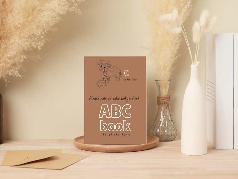 Op de afbeelding: Een bruine kaart met een zwart-witte illustratie van een koe en een kalf. De tekst op de kaart luidt "C is voor Cow. Help ons om baby's eerste ABC-boek te kleuren het leven op de boerderij."