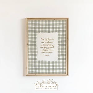 May include: A framed print featuring a handwritten prayer on textured paper. The background is a green and white gingham pattern. The frame is a light wood color. The text reads "May His favor be upon you...Amen."