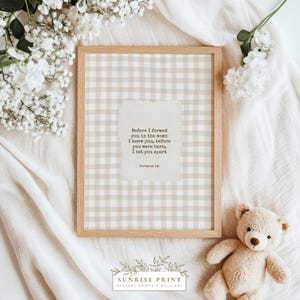 Puede incluir: Una impresión enmarcada con un marco de madera clara con un patrón de cuadros beige y blanco. La impresión incluye una cita de Jeremías 1:5 en un papel texturizado. Un osito de peluche y flores blancas están en la imagen.