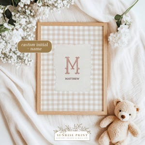 May include: A framed nursery print featuring a custom initial and name. The print has a light brown wooden frame and a gingham background. The central design element is a letter "M" in a dusty rose color, with the name "Matthew" below. A teddy bear is in the bottom right corner.