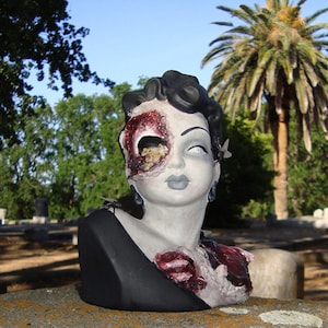 May include: A ceramic bust of a woman with a black dress and a gruesome, decaying face. The bust is painted with realistic details, including a missing eye and exposed flesh. The bust is displayed in a cemetery setting.