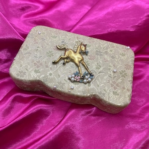May include: A vintage trinket box with a gold horse design and pearl accents. The box is covered in a floral patterned fabric and has a slightly curved shape.